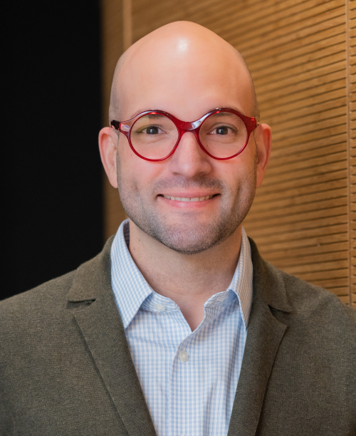 Dr. Rodriguez-Dias smiling with red glasses, blue collar shirt and brown jacket in front of a brown wall
